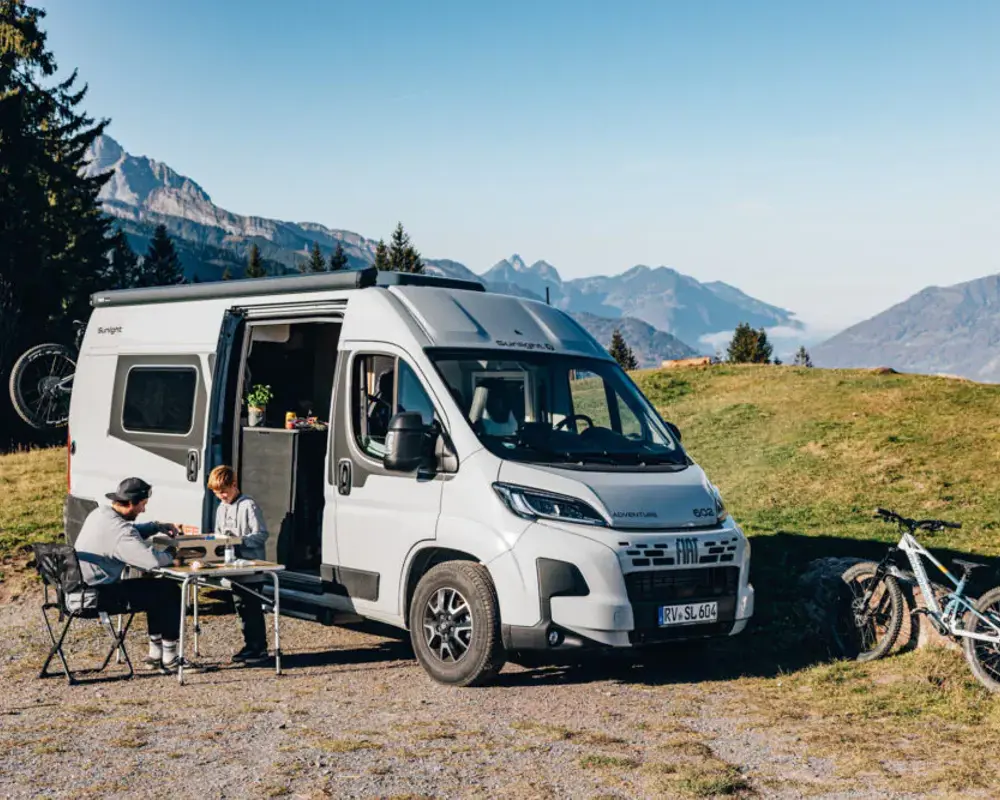 White camper van with people dining outdoors at a mountain campsite beside bicycles